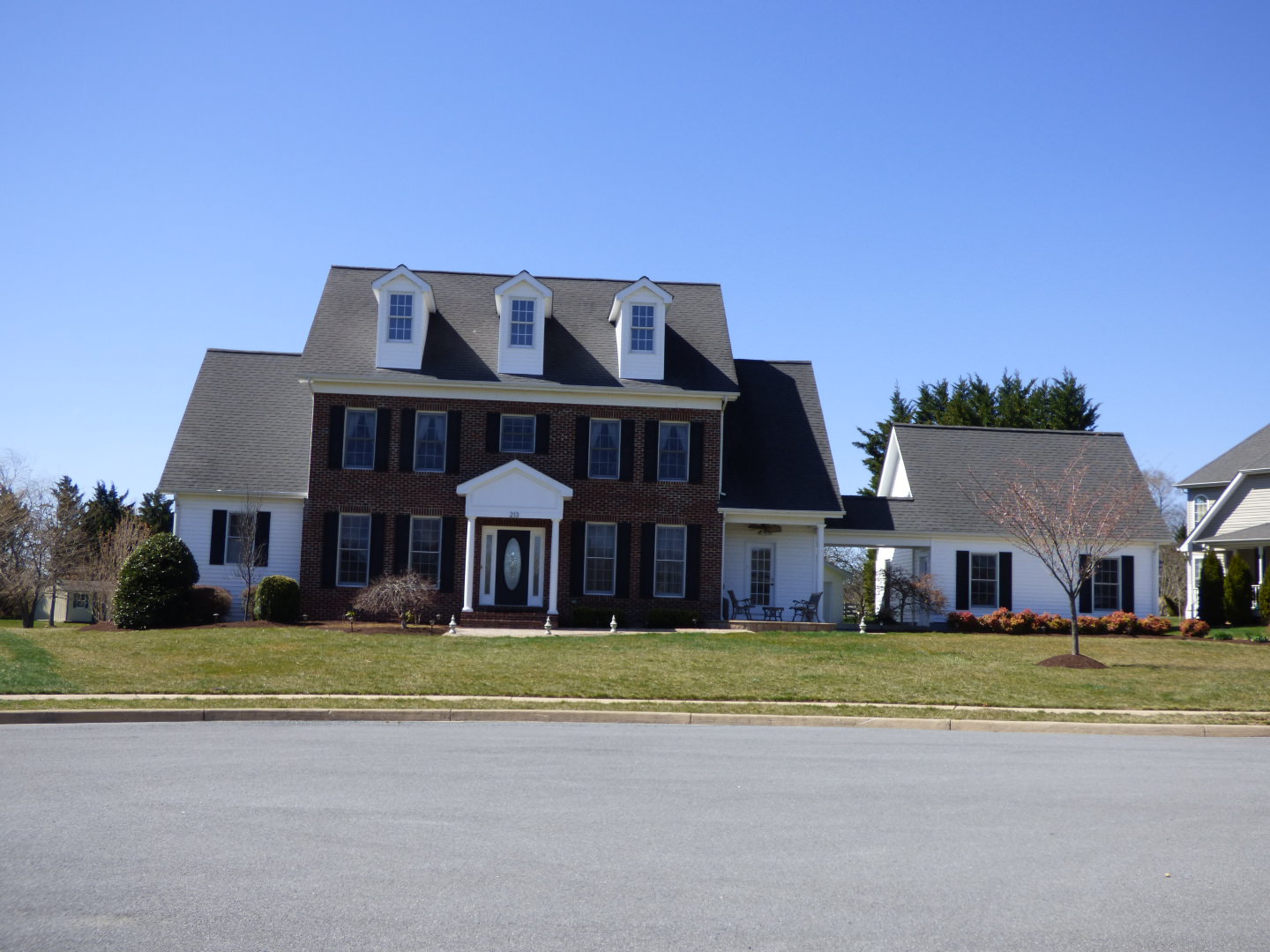 Large two-story brick house with white windows and black shutters, featuring a well-kept lawn and clear blue sky, conveying a serene suburban feel.