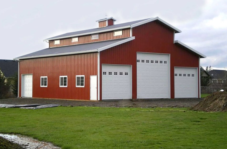 A large red barn with a gray metal roof and three white garage doors stands on a grassy lawn. It has several windows, creating a rustic, utilitarian feel.