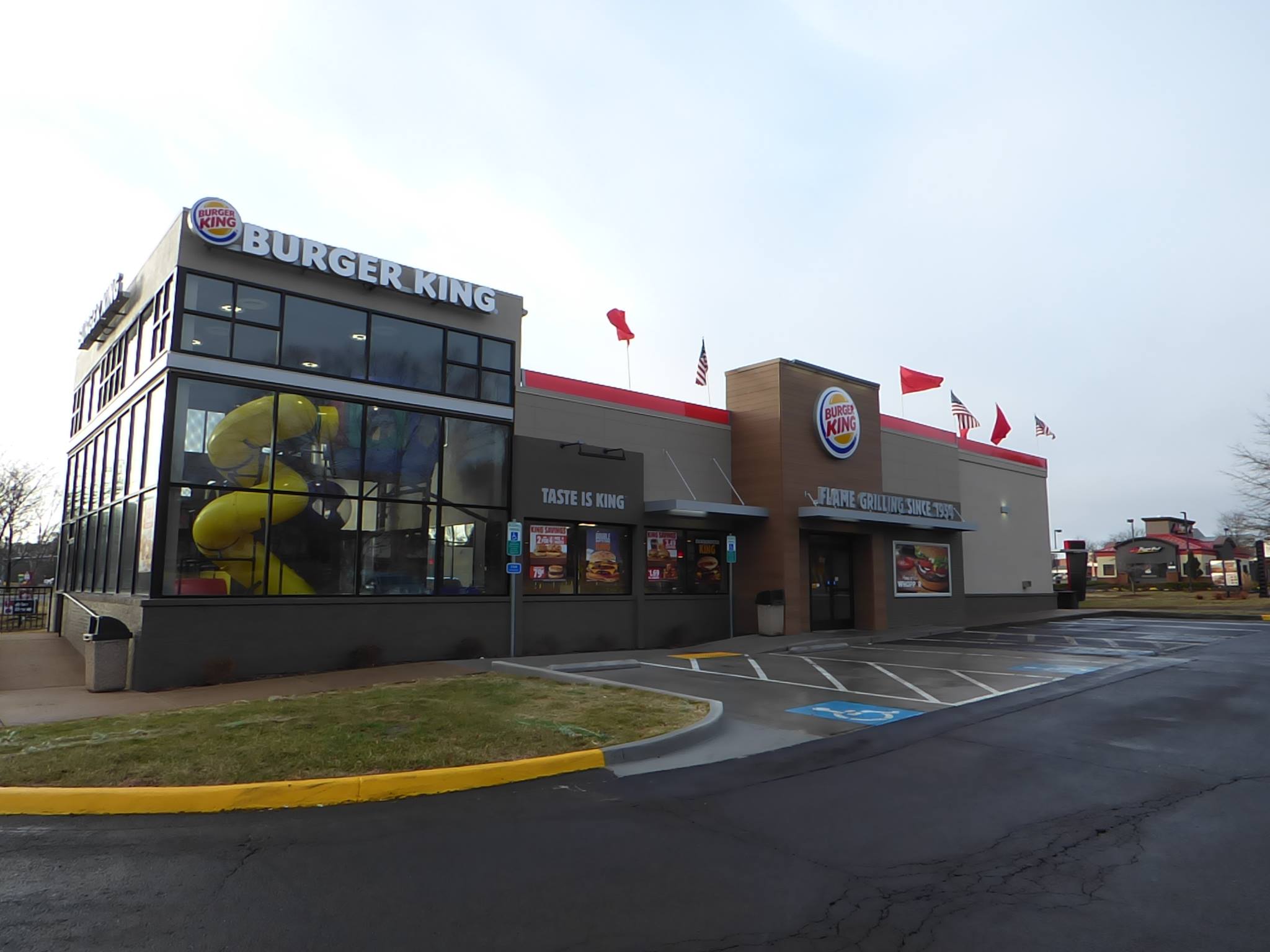 Burger King restaurant with a modern exterior, including bold signage, a visible indoor play area with a yellow slide, and an adjacent parking lot.