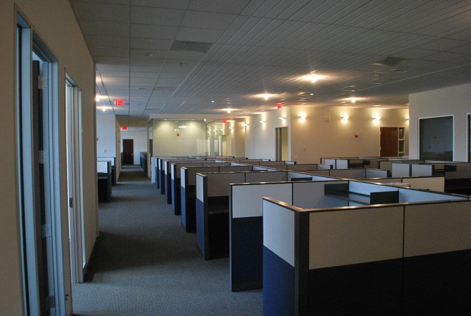 Dimly lit office corridor with rows of empty cubicles lined up, beige walls, gray carpet, and overhead fluorescent lights, creating a quiet atmosphere.