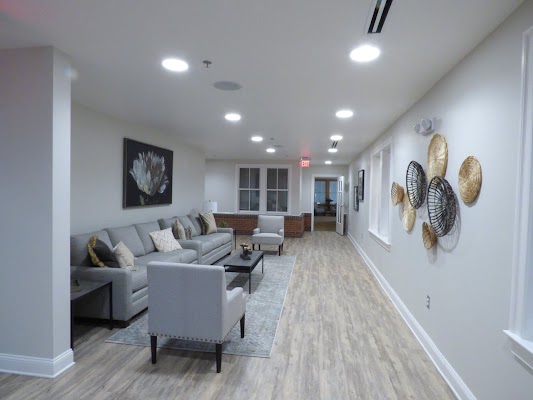 Sleek lounge with modern decor, featuring a gray sofa and armchairs, warm lighting, wall art, and wooden flooring, creating a cozy atmosphere.