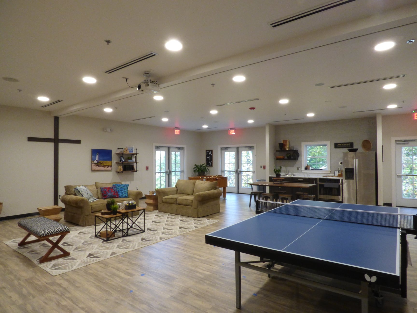 Spacious community room with cozy sofas, a rug, and a coffee table on the left. A ping pong table is on the right. Kitchen area and large windows in the background, creating a welcoming and interactive atmosphere.