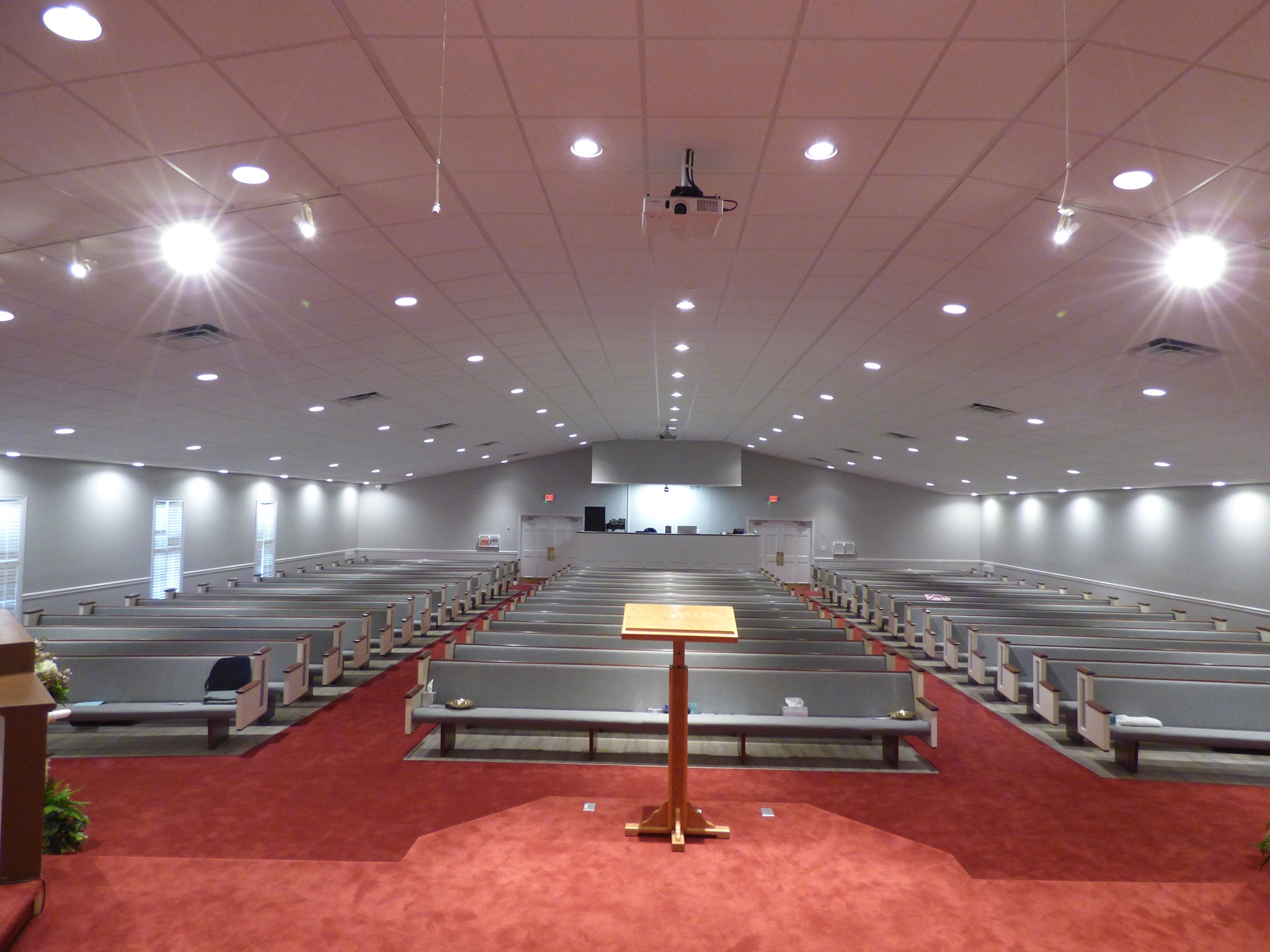 Spacious church interior with red carpet, central wooden podium, and rows of gray pews under bright ceiling lights, creating a serene and welcoming atmosphere.